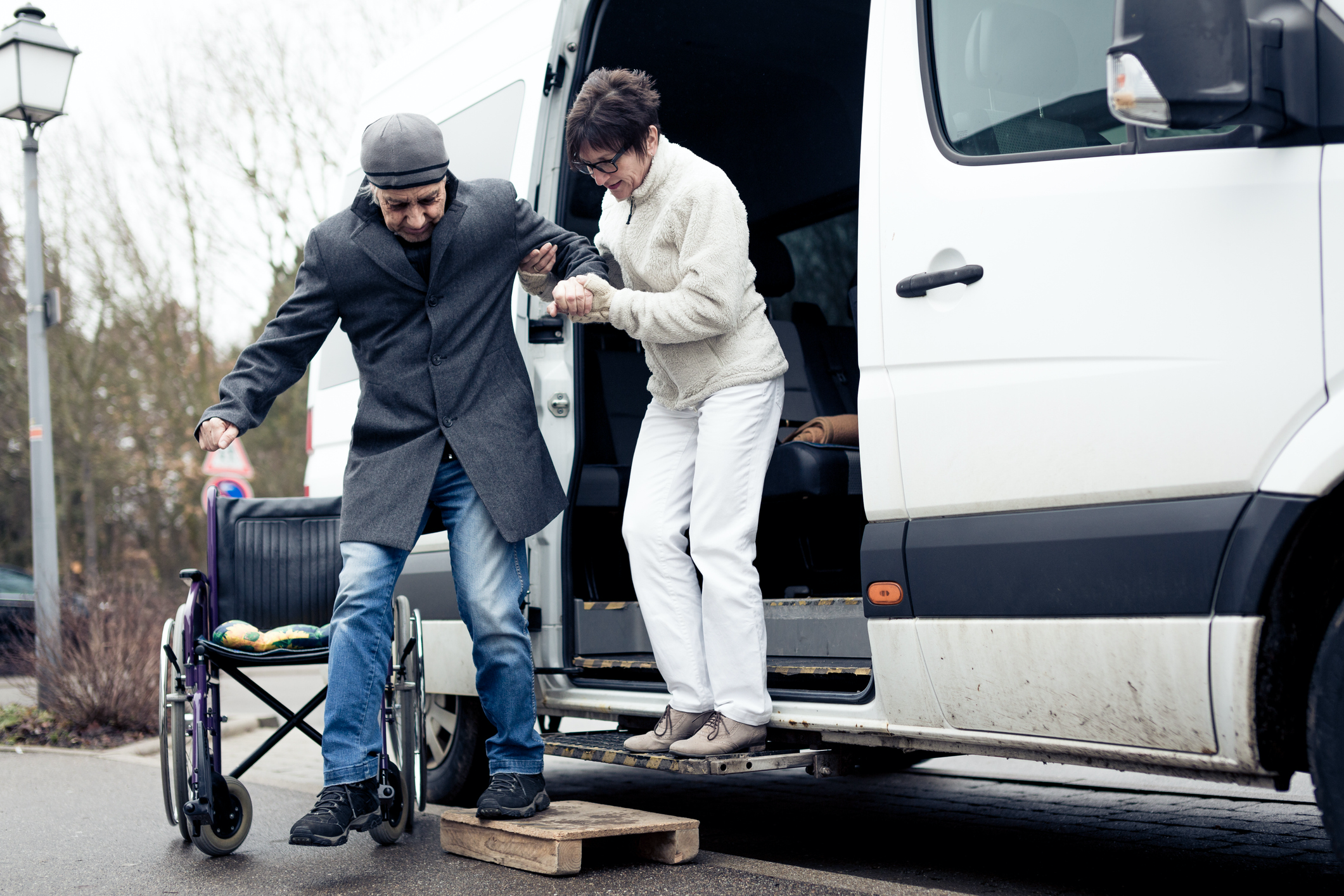 Woman Helps Elderly Man Out Of A Van And Into A Wheelchair