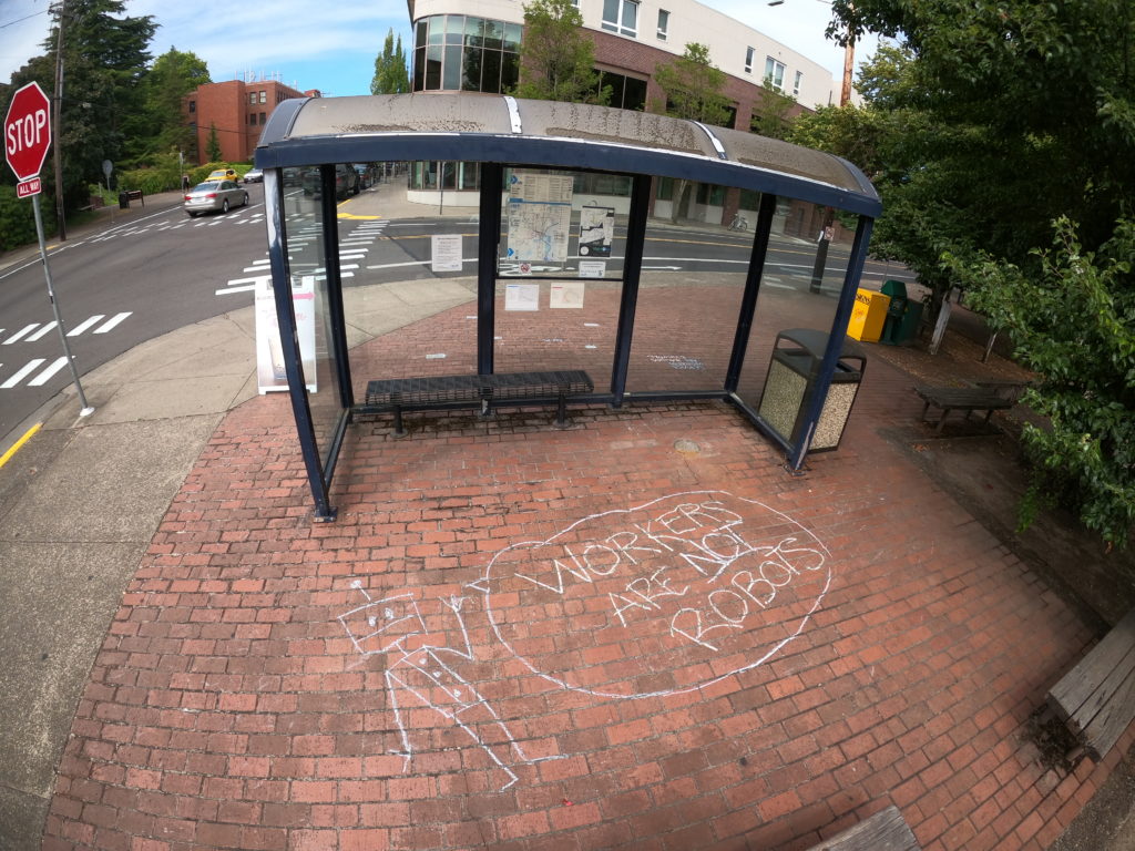 Photo of bus stop with chalk drawing on ground that says "workers are not robots"